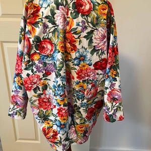 May include: A floral print blazer featuring a vibrant array of red, pink, yellow, blue, and purple flowers on a white background. The long-sleeved jacket is displayed on a wooden mannequin, showcasing its colorful design and tailored fit.
