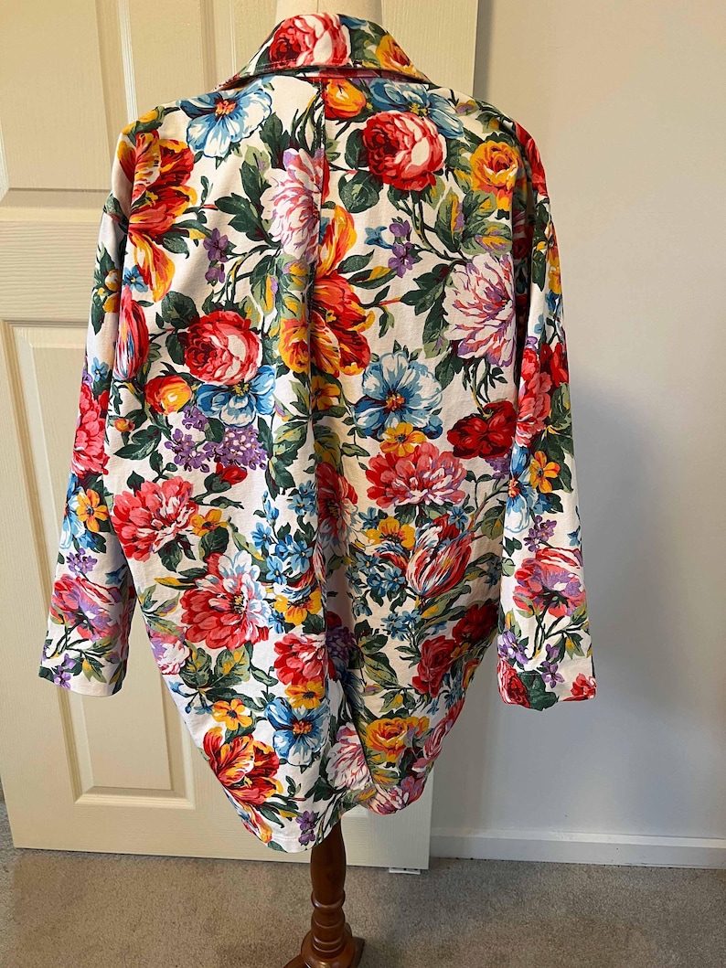 May include: A floral print blazer featuring a vibrant array of red, pink, yellow, blue, and purple flowers on a white background. The long-sleeved jacket is displayed on a wooden mannequin, showcasing its colorful design and tailored fit.