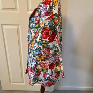 May include: A long-sleeved floral print shirt with a white background. The shirt features a vibrant pattern of red, yellow, blue, and purple flowers. The shirt is buttoned up and displayed on a wooden mannequin.
