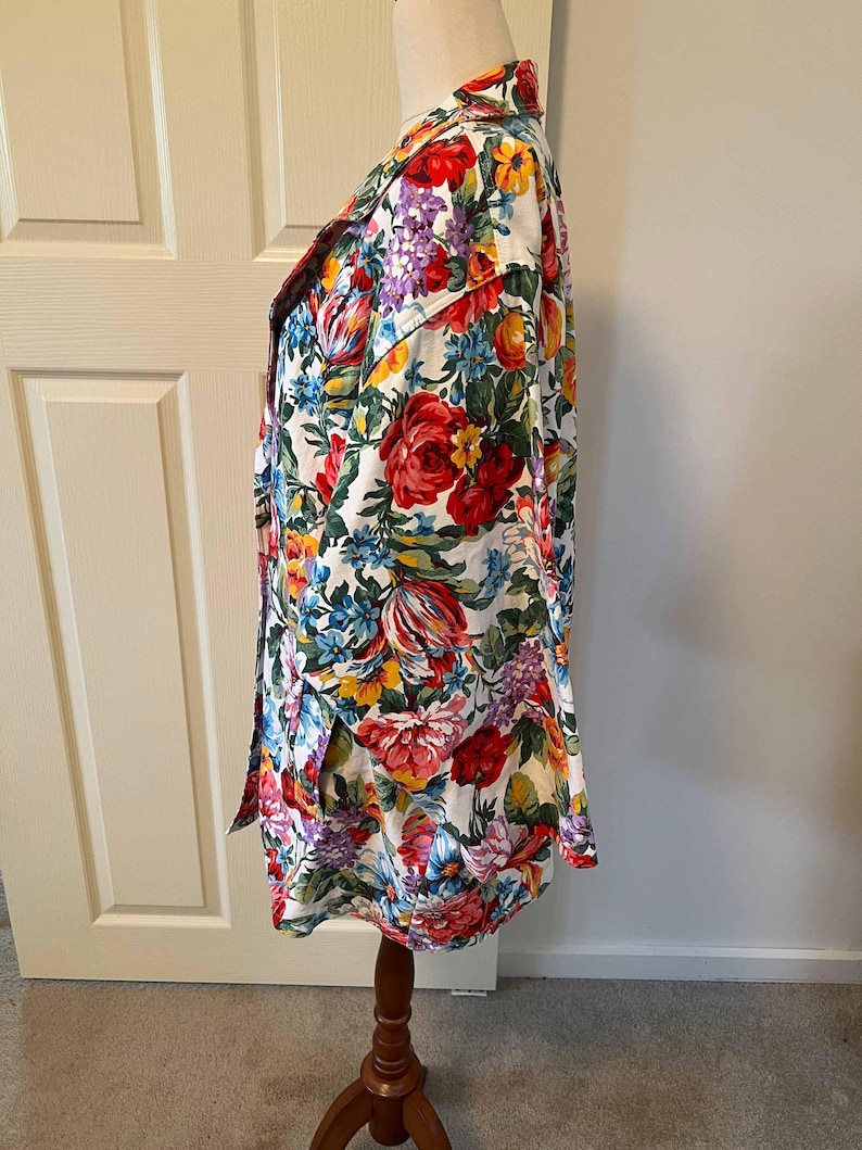 May include: A long-sleeved floral print shirt with a white background. The shirt features a vibrant pattern of red, yellow, blue, and purple flowers. The shirt is buttoned up and displayed on a wooden mannequin.