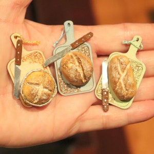 Può includere: Quattro taglieri in miniatura con piccoli coltelli e pagnotte di pane. I taglieri sono decorati con motivi floreali e hanno una gamma di colori blu chiaro, verde chiaro e marrone.