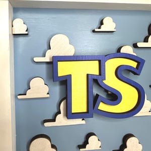 May include: A wooden wall sign featuring the letters TS in bright yellow with a thick blue outline. The letters are layered over a light blue background decorated with multiple small, white, three-dimensional cloud cutouts in a wooden frame.