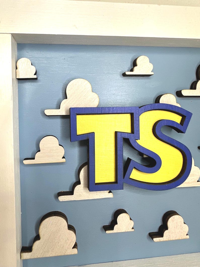 May include: A wooden wall sign featuring the letters TS in bright yellow with a thick blue outline. The letters are layered over a light blue background decorated with multiple small, white, three-dimensional cloud cutouts in a wooden frame.