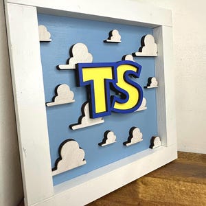 May include: A square wooden wall sign featuring a light blue background with multiple white, layered cloud cutouts. In the center, the letters TS are displayed in yellow with a thick blue outline, all mounted within a white wooden frame.