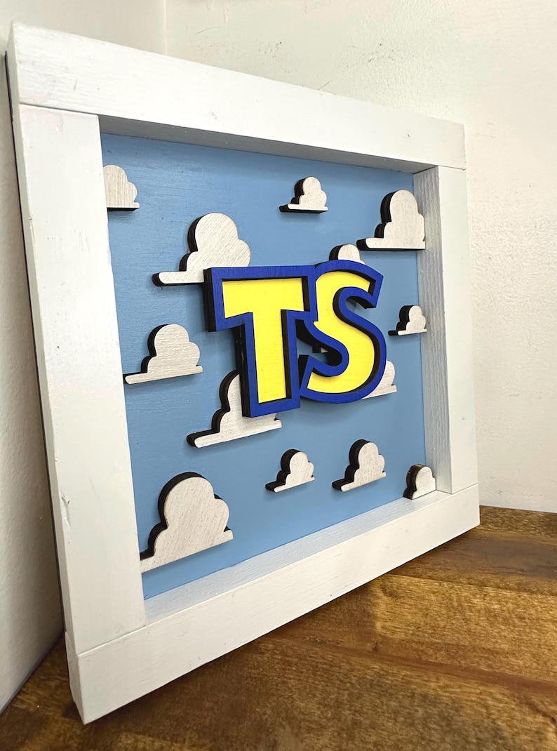 May include: A square wooden wall sign featuring a light blue background with multiple white, layered cloud cutouts. In the center, the letters TS are displayed in yellow with a thick blue outline, all mounted within a white wooden frame.