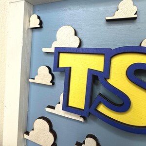 May include: A wooden wall sign featuring a light blue background with raised white cloud cutouts. The sign has a white wooden frame and displays large, bold yellow letters with a dark blue outline, partially showing the letters T and S.