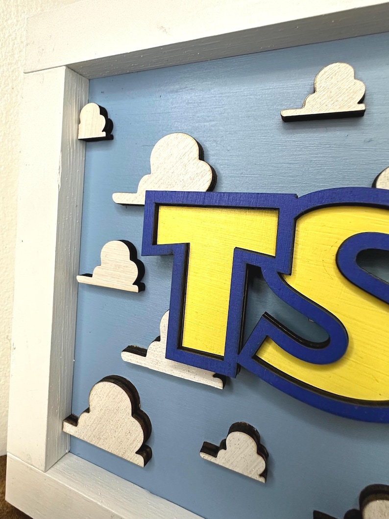 May include: A wooden wall sign featuring a light blue background with raised white cloud cutouts. The sign has a white wooden frame and displays large, bold yellow letters with a dark blue outline, partially showing the letters T and S.