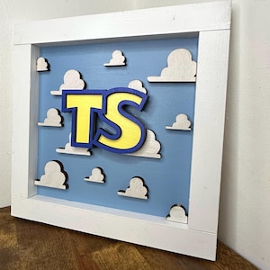 May include: A square wooden wall sign featuring a light blue background with white cloud cutouts. Centered on the sign are the yellow letters TS with a dark blue outline. The entire piece is enclosed in a thick, white wooden frame.