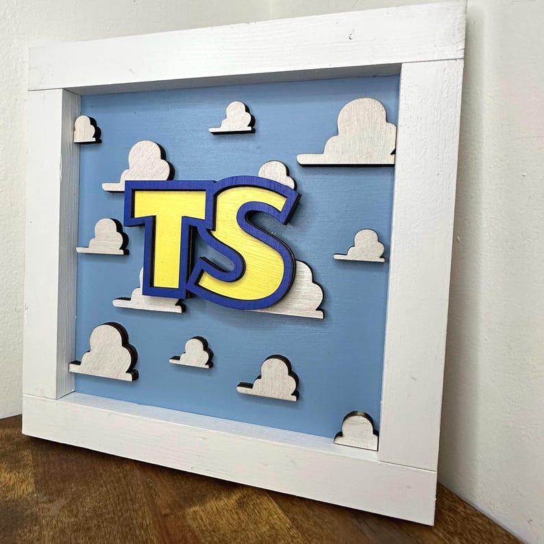 May include: A square wooden wall sign featuring a light blue background with white cloud cutouts. Centered on the sign are the yellow letters TS with a dark blue outline. The entire piece is enclosed in a thick, white wooden frame.