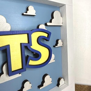 May include: A three-dimensional wall sign featuring the letters TS in bright yellow with a dark blue outline. The letters are mounted on a light blue background decorated with multiple white, cloud-shaped cutouts, all enclosed in a white frame.