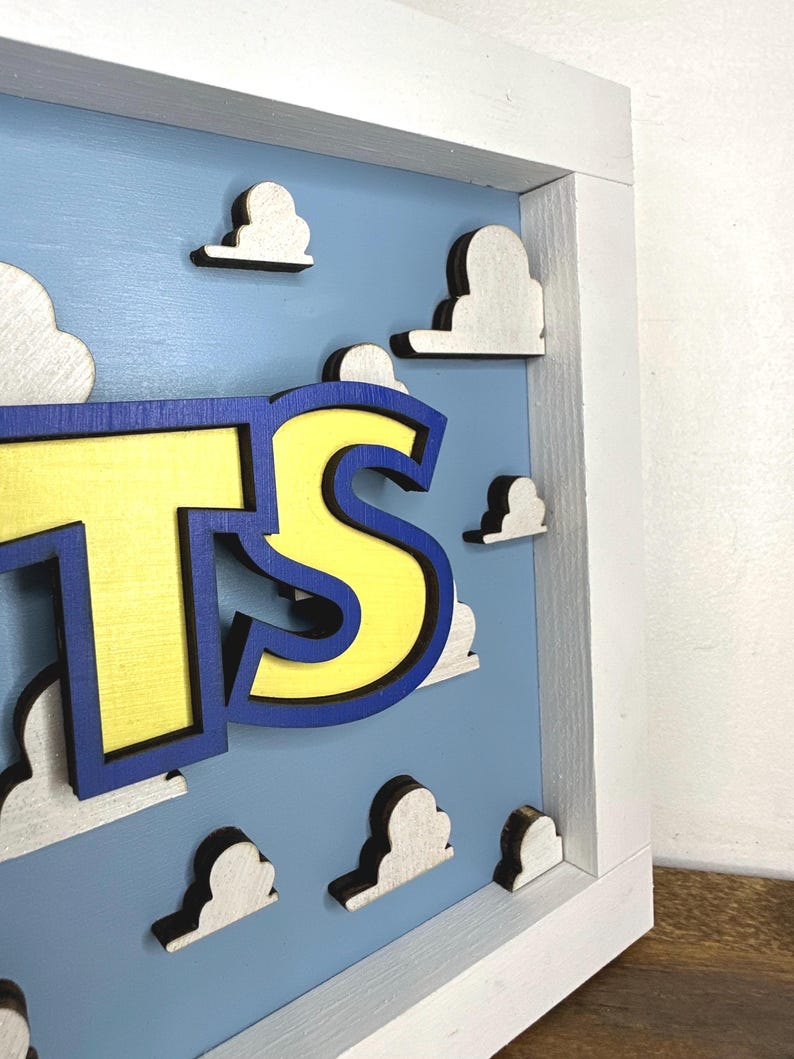 May include: A three-dimensional wall sign featuring the letters TS in bright yellow with a dark blue outline. The letters are mounted on a light blue background decorated with multiple white, cloud-shaped cutouts, all enclosed in a white frame.