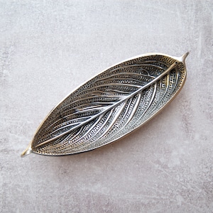 May include: A silver leaf-shaped dish with a textured surface. The dish is made of metal and has a slightly curved shape.