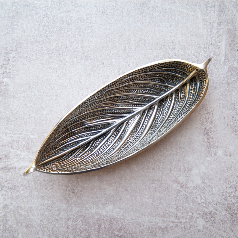 May include: A silver leaf-shaped dish with a textured surface. The dish is made of metal and has a slightly curved shape.