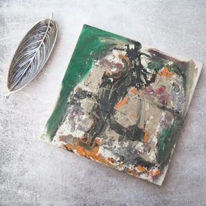 May include: Abstract painting on canvas with a green, black, white, orange, and gray color scheme. The painting has a textured surface and appears to be created with brushstrokes.
