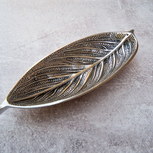 May include: A silver leaf-shaped dish with a detailed embossed design. The dish is made of metal and has a smooth, polished finish.