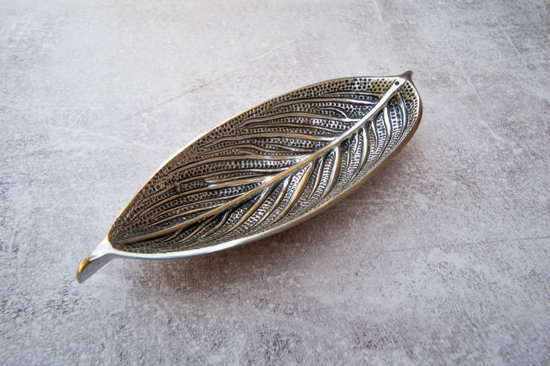 May include: A silver leaf-shaped dish with a detailed embossed design. The dish is made of metal and has a smooth, polished finish.