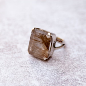May include: A silver ring with a large, rectangular, clear quartz stone with brown inclusions. The stone is set in a bezel setting.