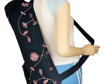 May include: A black yoga mat bag with a pink floral design. The bag is carried over the shoulder with a black strap. The bag is cylindrical and appears to be made of fabric. The bag is designed to carry a yoga mat.