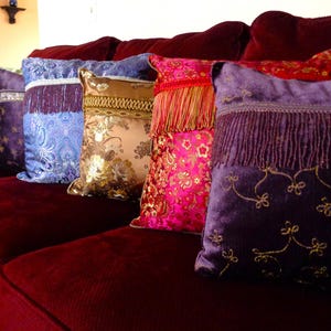 May include: A row of decorative throw pillows in various colors and patterns rests on a burgundy sofa. The pillows feature floral designs, fringe, and embroidered details. The color palette includes shades of purple, pink, gold, and blue.
