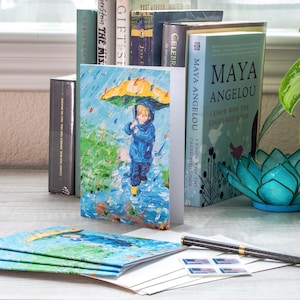 May include: Greeting cards featuring an impressionistic painting of a child in a blue raincoat and yellow umbrella in the rain. The cards are displayed with books, including "I Know Why the Caged Bird Sings" by Maya Angelou, and a pen.