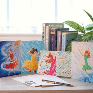 May include: Set of five greeting cards featuring colorful illustrations of a dancing figure in various poses. The cards showcase a vibrant palette of red, yellow, pink, and green against blue and white backgrounds. A pen and envelopes are also visible.