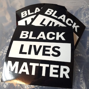 May include: A close-up of a stack of black and white "Black Lives Matter" stickers. The stickers are in a clear plastic container.