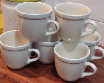 May include: Six white ceramic coffee mugs with a brown rim. The mugs are stacked on top of each other on a wooden surface.