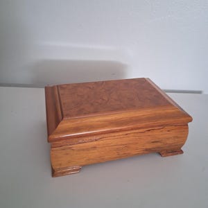May include: A rectangular wooden box with a polished finish, featuring a distinct burl wood grain pattern on the lid and a smooth, lighter-toned wood grain on the sides. The box has a beveled edge around the top and sits on four small feet.