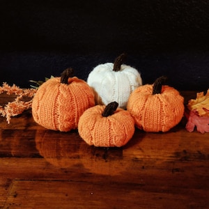 May include: Four knitted pumpkins, three orange and one white, on a wooden surface. The pumpkins are decorated with a cable knit pattern.