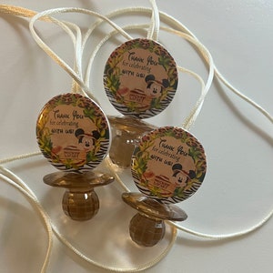 May include: Three brown pacifiers with round, decorative tops and attached cream-colored cords. Each pacifier has a circular tag with the text "Thank You for celebrating with us!" and a Mickey Mouse graphic, with a safari theme.