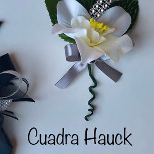 Pu&ograve; includere: Un'occhiello floreale bianco e crema con un nastro argentato e un accento di strass. L'occhiello ha foglie verdi e uno stelo verde riccio. Il testo "Cuadra Hauck Creation's" &egrave; scritto sotto.