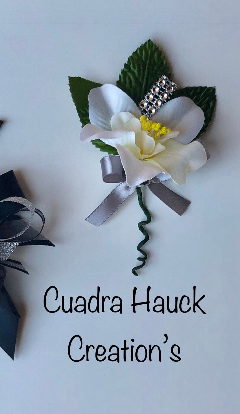 Pu&ograve; includere: Un'occhiello floreale bianco e crema con un nastro argentato e un accento di strass. L'occhiello ha foglie verdi e uno stelo verde riccio. Il testo "Cuadra Hauck Creation's" &egrave; scritto sotto.
