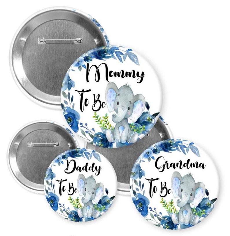 May include: Baby shower badges featuring a blue floral design and a grey elephant illustration. The badges read "Mommy To Be", "Daddy To Be", and "Grandma To Be". The badges are round with a pin fastening on the back.