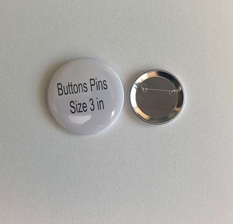 May include: Two white, round buttons with a pin back. One button has the text "Buttons Pins Size 3 in" printed in black. The other button shows the silver pin back. The buttons are on a white background.