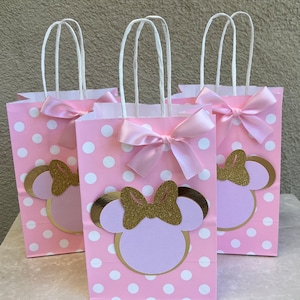 May include: Three pink gift bags with white polka dots, featuring a Minnie Mouse design. Each bag has a gold glitter bow, a light pink bow, and white handles. The bags are arranged on a light-coloured surface.