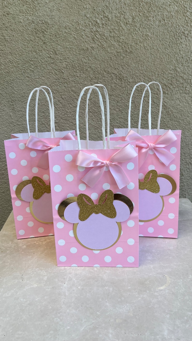 May include: Three pink gift bags with white polka dots, featuring a Minnie Mouse design. Each bag has a gold glitter bow, a light pink bow, and white handles. The bags are arranged on a light-coloured surface.