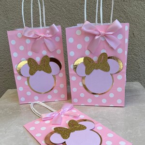 May include: Pink polka dot gift bags with white handles, decorated with a gold glitter bow and a Minnie Mouse design. Each bag has a pink satin bow. Ideal for party favours or gifts.