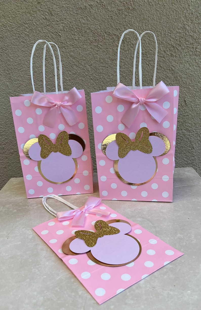 May include: Pink polka dot gift bags with white handles, decorated with a gold glitter bow and a Minnie Mouse design. Each bag has a pink satin bow. Ideal for party favours or gifts.