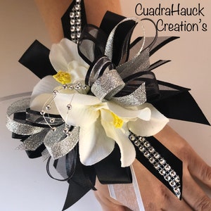 Pu&ograve; includere: Un corsage da polso con fiori bianchi, centri gialli e nastri neri e argentati. Impreziosito da strass e perline argentate. Il testo "CuadraHauck Creation's" &egrave; visibile.
