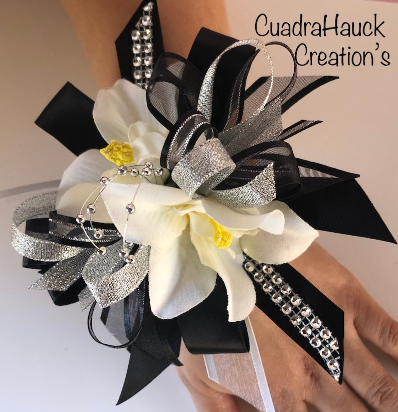 Pu&ograve; includere: Un corsage da polso con fiori bianchi, centri gialli e nastri neri e argentati. Impreziosito da strass e perline argentate. Il testo "CuadraHauck Creation's" &egrave; visibile.