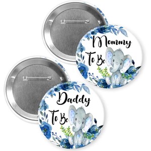 May include: Two round baby shower badges with a blue floral design and a grey elephant illustration. One badge reads "Mommy To Be" and the other reads "Daddy To Be". The badges have a silver pin back.