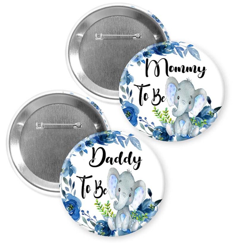 May include: Two round baby shower badges with a blue floral design and a grey elephant illustration. One badge reads "Mommy To Be" and the other reads "Daddy To Be". The badges have a silver pin back.