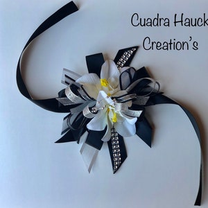 Pu&ograve; includere: Un fiocco floreale decorativo con nastri bianchi, neri e argentati. Il fiocco presenta un fiore bianco con accenti gialli e abbellimenti di strass. Il testo "Cuadra Hauck Creation's" &egrave; visibile.