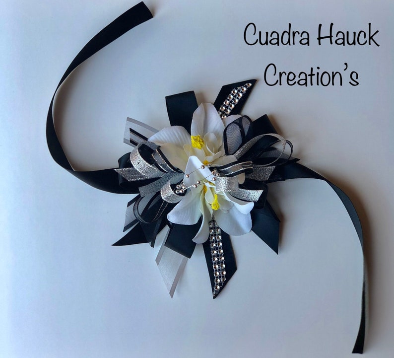 Pu&ograve; includere: Un fiocco floreale decorativo con nastri bianchi, neri e argentati. Il fiocco presenta un fiore bianco con accenti gialli e abbellimenti di strass. Il testo "Cuadra Hauck Creation's" &egrave; visibile.