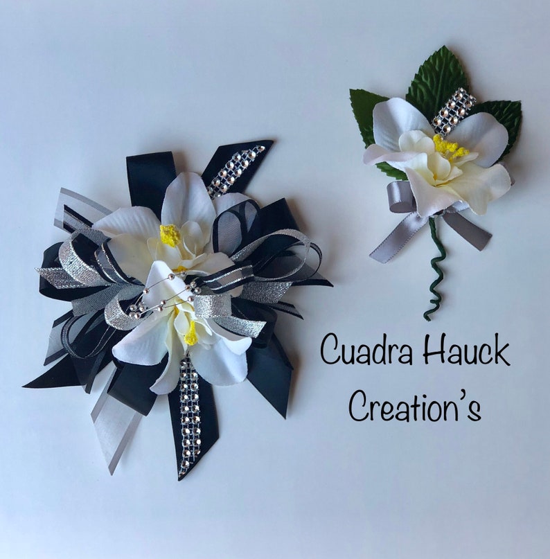Pu&ograve; includere: Due accessori floreali: un grande fiocco e un'occhiello. Il fiocco presenta fiori bianchi, nastri neri e argentati e strass. L'occhiello ha un fiore bianco, foglie verdi e un nastro argentato. Il testo "Cuadra Hauck Creation's" &egrave; visibile.