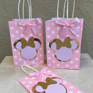 May include: Pink polka dot gift bags with white handles, featuring a Minnie Mouse design. Each bag has a gold glitter bow and a pink satin bow. Ideal for a themed party or celebration.