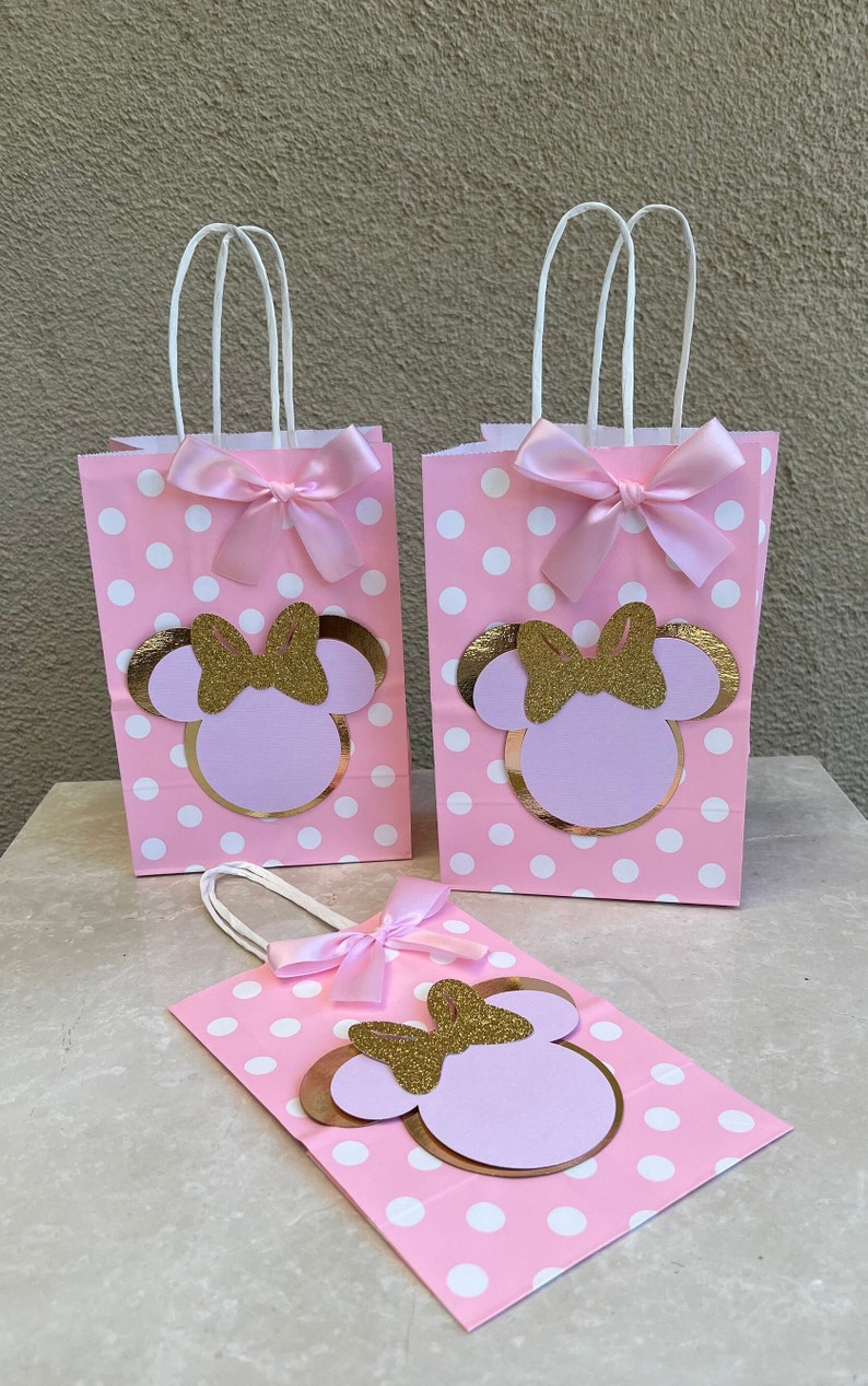 May include: Pink polka dot gift bags with white handles, featuring a Minnie Mouse design. Each bag has a gold glitter bow and a pink satin bow. Ideal for a themed party or celebration.