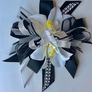 Pu&ograve; includere: Un fiocco floreale decorativo con fiori di tessuto bianco con centri gialli, accentuato da nastri neri, argentati e grigi. Il fiocco &egrave; impreziosito da un filo di perline argentate e un nastro tempestato di strass.