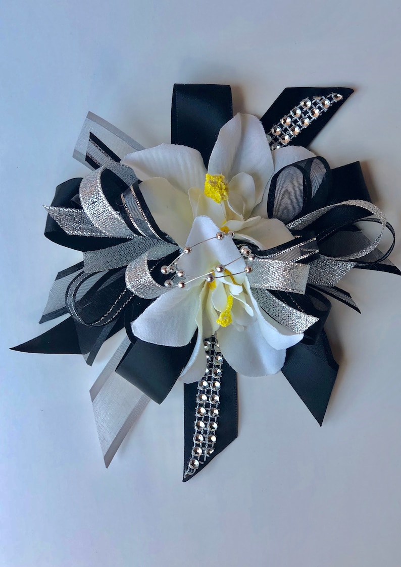 Pu&ograve; includere: Un fiocco floreale decorativo con fiori di tessuto bianco con centri gialli, accentuato da nastri neri, argentati e grigi. Il fiocco &egrave; impreziosito da un filo di perline argentate e un nastro tempestato di strass.