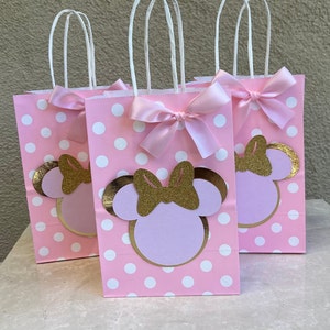 May include: Three pink polka dot gift bags with white handles and pink bows. Each bag features a Minnie Mouse design with a gold glitter bow and gold outline. The bags are for a party.